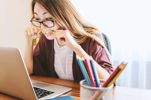 Frau mit langen, braunen Haaren und dunkler Brille sitzt an einem Schreibtisch vor dem aufgeklappten Laptop und beißt frustriert in einen gelben Stift