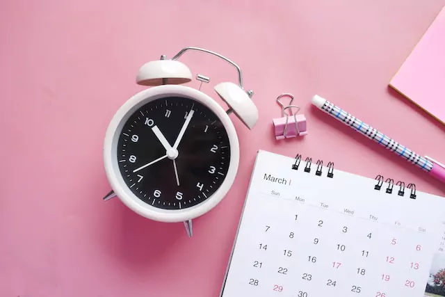 auf pinkem Hintergrund liegt ein Wecker, ein Kalender und ein Stift